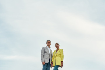 Just happy together. Elegant and lovely middle-aged couple holding hands and looking away while standing against blue sky