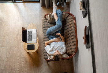 Woman use a smartphone while lying on a sofa with a laptop and ereader on a table. Studying online,...
