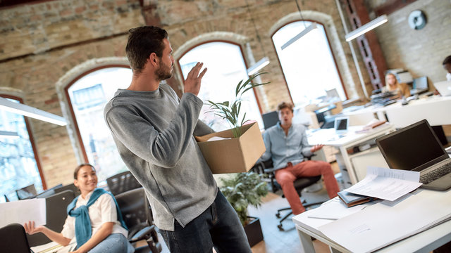 Follow Your Dreams, Quit Your Job. Handsome Man In Casual Wear Holding Box With Personal Things And Saying Good Bye To His Ex-colleagues While Leaving Modern Office