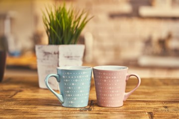 Two cups on wooden table, plant and kitchen in the background.