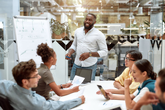 Great Work. Young Afro American Businessman Pointing At White Blackboard And Smiling While Working With Multicultural Team In The Creative Office