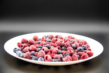 A Berry mix  from frozen raspberries and blueberries on the white plate. A Frozen Berries in black background.  A sweet background with frozen raspberries and blueberries