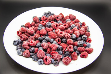 A Berry mix  from frozen raspberries and blueberries on the white plate. A Frozen Berries in black background.  A sweet background with frozen raspberries and blueberries