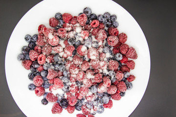 A Berry mix in sugar from frozen raspberries and blueberries on the white plate. A Frozen Berries with Sugar.  A sweet background with frozen raspberries and blueberries