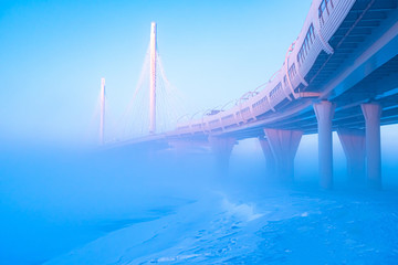 The bridge in the winter. Road bridge in the blue fog. Winter road landscape. Freeway on supports. Road infrastructure. Traffic. Winter landscape with cable-stayed bridge.