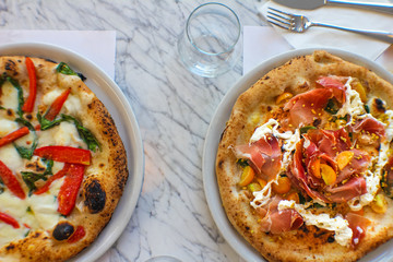 top view of pizza with corbarino yellow tomato, black Casertano pork ham, chopped pistachio and buffalo stracciatella and another one with sliced San Marzano tomato