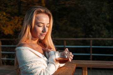 Charming woman is drinking hot tea in the cold outdoor from a glass cup. Happy european blonde girl is keeping warm with hot tea in the cold autumn city park. Catch a cold...