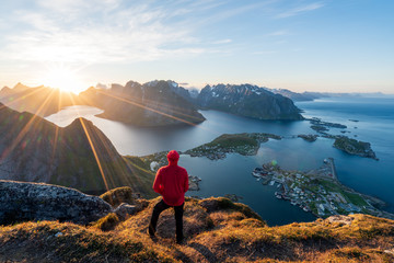 Hiker in red jacket are standing on top of the famous Reinebringen overlooking Reine and Hamnoy in the Lofoten islands during the midnight sun. Active, landscape and climbing concept. - obrazy, fototapety, plakaty