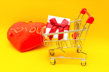 gift box in shopping cart on yellow background next to red hearts, copy space. Concept of gifts,...