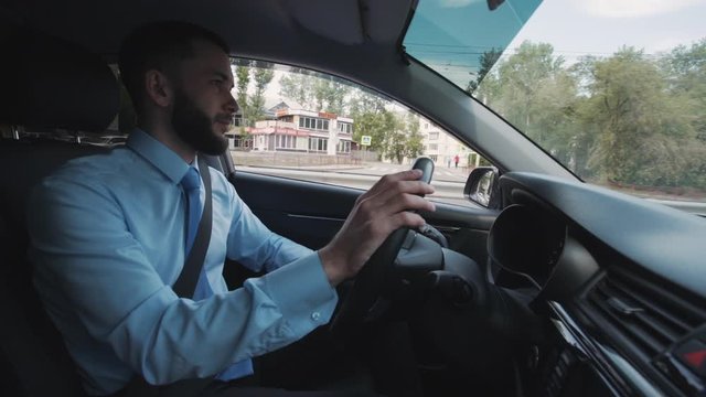 Young american businessman driving car, watching front sitting in auto cab. Confident male driver holding steering wheel in hand, riding in city street. Bearded man wearing shirt and tie. Concept