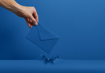 Male hand holding an envelope with a letter through torn classic blue color paper background. The...