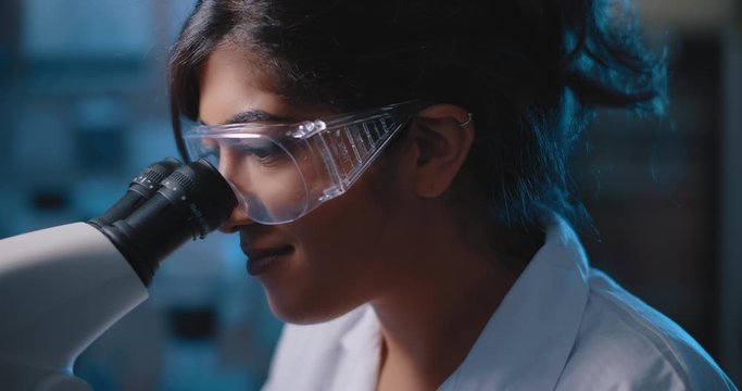 Female research scientist looking into microscope, wearing safety glasses.Blue lighting in a dark lab room.Close up, slider, shot with BMPCC 4K.Biochemistry, pharmaceutical medicine, science concept