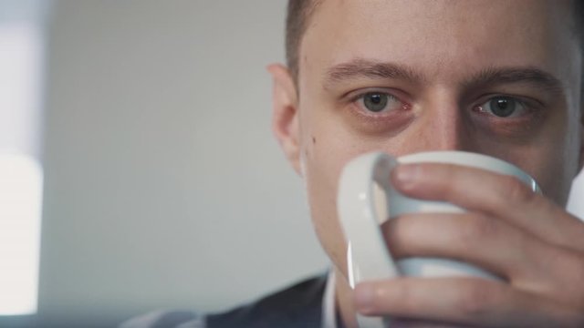 Happy American Businessman Drinking Coffee On White Background In Office Room. Facial View Of Smiling Man Is Sipping Beverage, Holding Cup In Hand And Watching Front In Interior. Concept: Breakfast