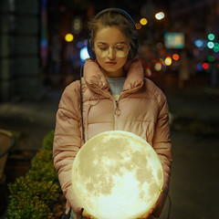 Naklejka premium Romantic portrait of a young girl on the street of the evening city with the moon in her hands