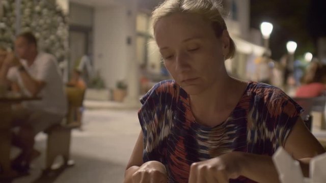 Young blond woman sits in an outdoor restaurant at night; cuts and eats pizza with a knife and fork.