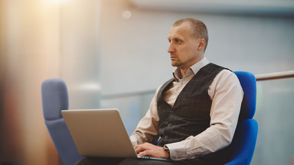 Portrait of a mature confident man entrepreneur sitting on a classic blue armchair with his laptop and pensively looking in front; a caucasian businessman with a netbook in an office open-space area