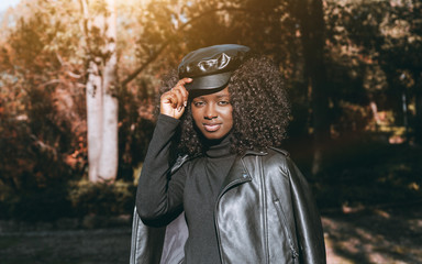 An outdoor portrait of a cute young curly-hair African female in a leather jacket, peaked cap, black pullover, with park trees in a defocused background; a charming black Guinean girl outdoors