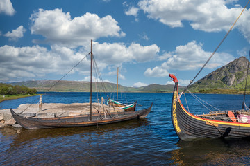 Viking warrior ancient  wooden long ships in harbour with blue cloudy sky and green mountain in the background. Vikings, war equipment and history concept.