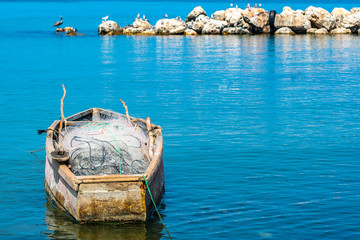 Empty old wooden fishing boat with net and rods, docked & floating in sea water. Coastal ocean view with birds perched on large sea rocks/ boulders in the background. Sunny summer tropical island day.