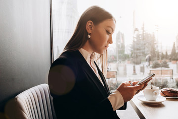 a beautiful girl wearing in office clothes and writing on the phone  in the cafe with coffe and cake on the table on the sunny day