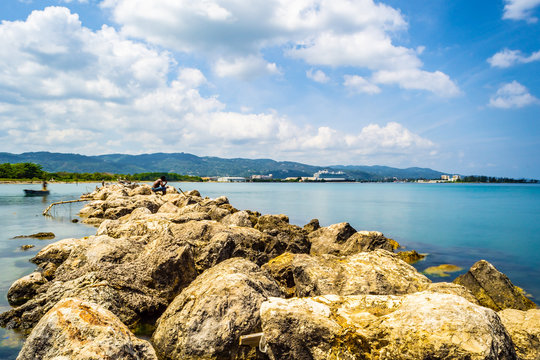 Scenic ocean water view setting from the north coast of Montego Bay, Jamaica, with large sea boulders and cruise ship and mountains in background. Black Jamaican man wearing no shirt sitting on rocks. © Debbie Ann Powell