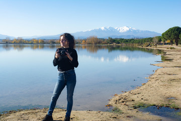 Fototapeta na wymiar Girl with curly hair takes pictures in a lake with the mountains in the background.Lake of Villeneuve-de-la-Raho (France) overlooking the Pyrenees and the Canigo. Concept: Landscape photography