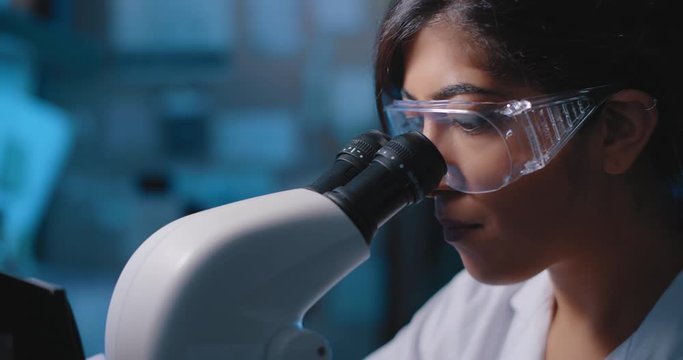 Female Research Scientist Looking Into Microscope, Wearing Safety Glasses.Blue Lighting In A Dark Lab Room.Close Up, Slider, Shot With BMPCC 4K.Biochemistry, Pharmaceutical Medicine, Science Concept