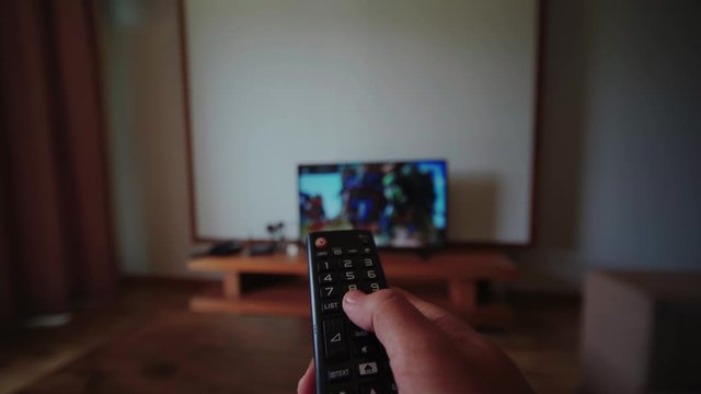 Hand holding tv remote control, changing channel on television, watching movie at living room in house, chilling alone.