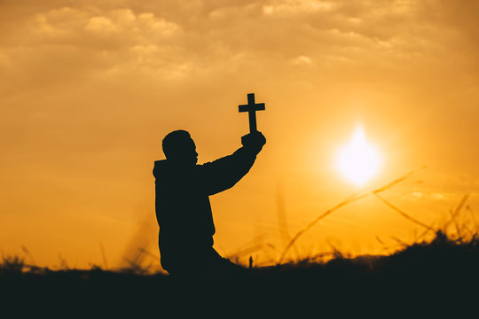 Christian man sitting praying with holding christian cross at sunset background. christian silhouette concept.