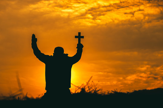 Christian man sitting lift hands and praying to God with christian cross at sunset background. christian silhouette concept.