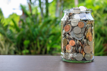 Many coins are glass jar for saving money on table and nature background.