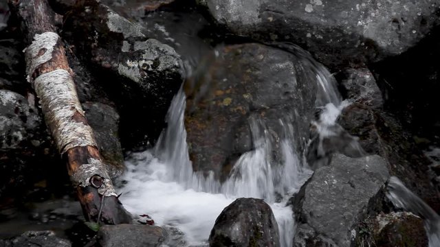 A small mountain waterfall cascades over rocks and boulders with water splashing down in this seamless video loop