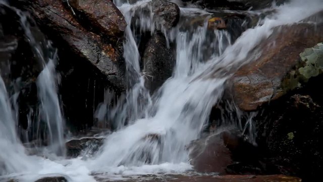 A small mountain waterfall cascades over rocks and boulders with water splashing down in this seamless video loop