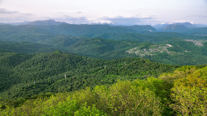 Green hills covered with dense forest and high snowy mountains in the distance. Beautiful sunset in a hilly valley with villages and fog in the lowlands.