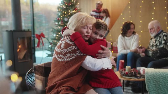 On Christmas days, cute happy grandson giving a holiday present to grandmother. Cheerful lovable grandpa and kid exchanging gigts hugging together joy celebrating at home.