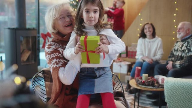 Adorable little girl holding a present approaching to grandma sitting on her knees exchanging family gift boxes on Christmas Eve. Happy family on boxing day. Winter holidays.