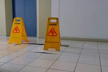 Closeup buckled tiles on walkway in front of elevator, there are two caution sign used for warning...