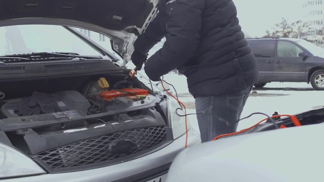 Man connects jumper clamp cables to automotive battery post terminal. Hood or bonnet raised up in winter daytime in parking lot of yard. Malfunction or problem with auto. Part 3