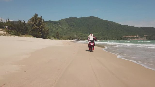 Santa Claus on the seashore rides a bike with a bag of gifts.