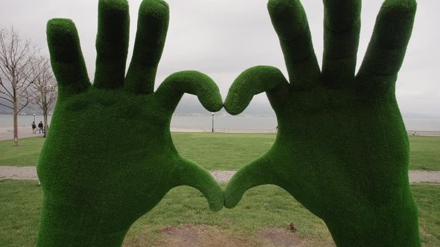 big topiary grass hands making heart sign shape in the park by sea. sculpted green foliage. greenery landscape. cloudy weather. concept lawn, passers-by, people