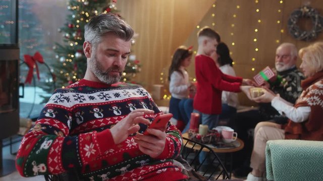 Busy bearded man using smartphone on family Christmas holiday celebration. Upset father businessman working online while his kids and parents exchanging gifts at boxing day. Sad Christmas.