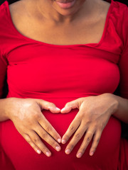 A young African American mixed race woman wearing a bright red dress looks down and holds her hands on her pregnant belly forming her fingers in the shape of a heart making a beautiful maternity photo