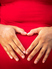 A young African American mixed race or ethnicity woman wearing a bright red dress holds her hands on her pregnant belly forming her fingers in the shape of a heart making a beautiful maternity photo.