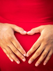 A young African American mixed race or ethnicity woman wearing a bright red dress holds her hands on her pregnant belly forming her fingers in the shape of a heart making a beautiful maternity photo.