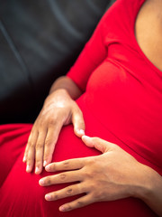 A young African American mixed race or ethnicity woman wearing a bright red dress holds her hands on her pregnant belly forming her fingers in the shape of a heart for a maternity pose.