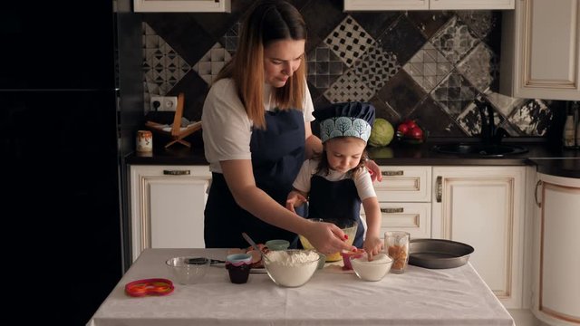 Cute young mother and little daughter cook pastries together at home in the kitchen. Mom with a child in identical aprons eat sugar licking a finger.
