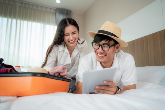 Happy couple packing suitcase on bed in bedroom and lying and looking digital tablet for search and planning travel trip online. Asian backpacker travel lifestyle concept..