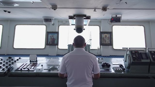 back view of captain of the cargo ship standing by the dashboard and steering wheel. russian man controls transport. concept of business, job, icon, cabin, deck.