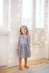 The girl sits near the window decorated with a garland for the holiday, the room is decorated for the new year and Christmas