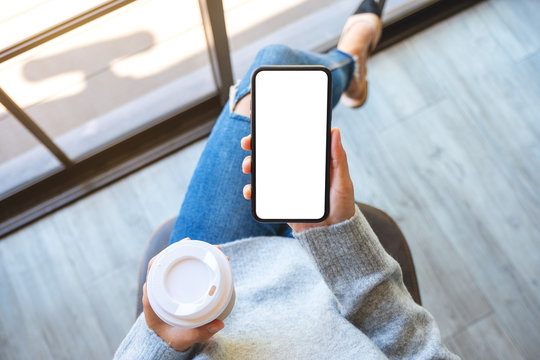 Top view mockup image of a woman holding a black mobile phone with blank white desktop screen with coffee cup